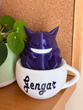 Figura decorativa de Gengar dormido dentro de una taza blanca con detalles morados